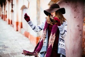 Ragazza che indossa un cardigan sui toni dell'azzurro con motivi invernali abbinato ad una maxi sciarpa e cappello a tesa larga nelle fantasie del vinaccia e marrone