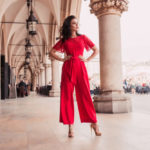 Immagine ambientata in un porticato arricchito da colonne. La ragazza indossa una tuta color corallo con pantaloni a palazzo, fusciacca in vita dello stesso colore e maniche corte con inserti e finiture in pizzo. L'outfit è accompagnato da sandali a tacco alto color oro con motivi intrecciati sul fronte.