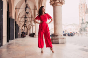 Immagine ambientata in un porticato arricchito da colonne. La ragazza indossa una tuta color corallo con pantaloni a palazzo, fusciacca in vita dello stesso colore e maniche corte con inserti e finiture in pizzo. L'outfit è accompagnato da sandali a tacco alto color oro con motivi intrecciati sul fronte.