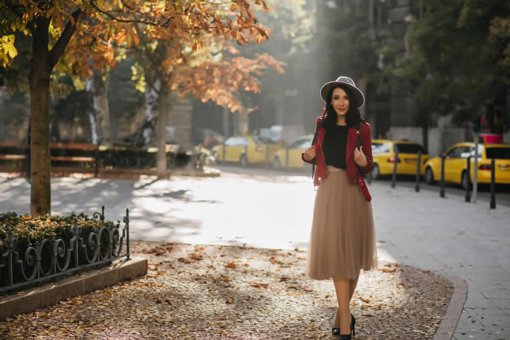 Ragazza in un parco in autunno. Indossa una gonna in tulle sotto il ginocchio abbinata ad un sottogiacca nero e giacca rosso ciliegia. Completano l'outfit cappello modello borsalino azzurro polvere e décolleté nere.