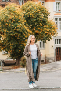 Ragazza a figura intera con sfondo di villa e albero autunnale. Indossa un cardigan lungo color cammello abbinato a jeans blu a sigaretta e t shirt bianca