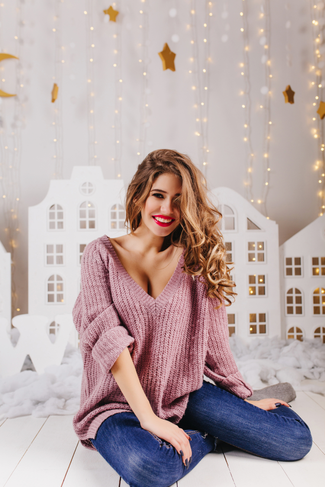 Immagine con sfondo invernale bianco e oro. Ragazza bionda seduta che indossa un maxi pull rosa con scollo a V e jeans blu