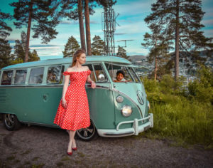 Immagine di una ragazza appoggiata ad un vecchio furgone Wolkwagen anni 50 color verde salvia. La ragazza indossa un abito rosso a pois bianchi senza spalline modello a campana anni 50, in abbinamento a décolleté a tacco alto di colore rosso