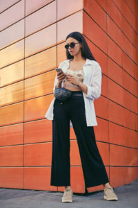 Ragazza che indossa pantaloni a palazzo neri con top corto fantasia e lunga camicia bianca in abbinamento. Accessori: piccolo marsupio in pelle nera trapuntata e sneakers color corda