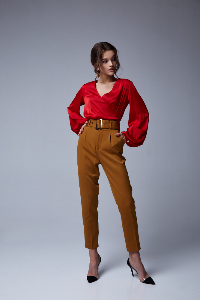 Immagine di ragazza a figura intera che indossa pantaloni color cognac con cintura alta e camicetta in raso rossa con maniche a sbuffo e scollo a V. Scarpe décolleté nere con tacco alto e fascia oro.