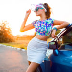 Ragazza al tramonto appoggiata ad un auto che indossa mini gonna bianca e top corto multicolor