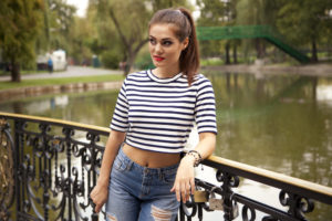 Immagine di ragazza su un ponte con sfondo di un fiume. Indossa jeans azzurro chiaro con tagli sul davanti in abbinamento ad un crop top modello Marina con righe orizzontali bianche e blu
