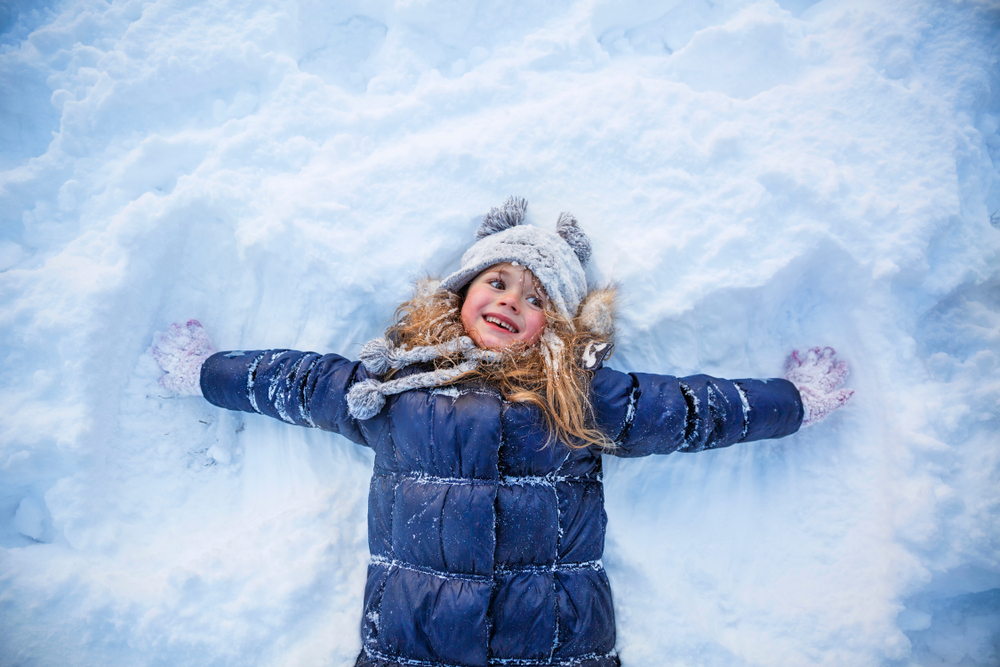 Bimba distesa nella neve. Indossa un piumino di colore blu e grande cappello con pon pon grigio