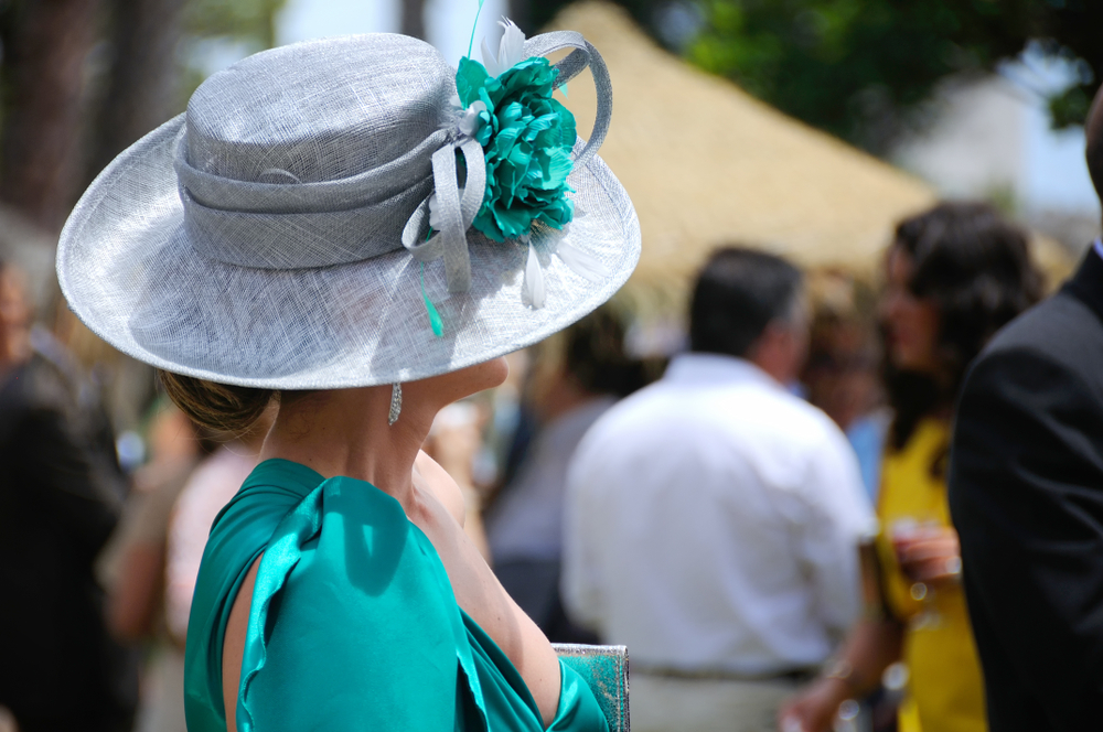 Donna vista di profilo. Indossa un abito turchese da cerimonie completato da un cappellino grigio perla e fiore turchese